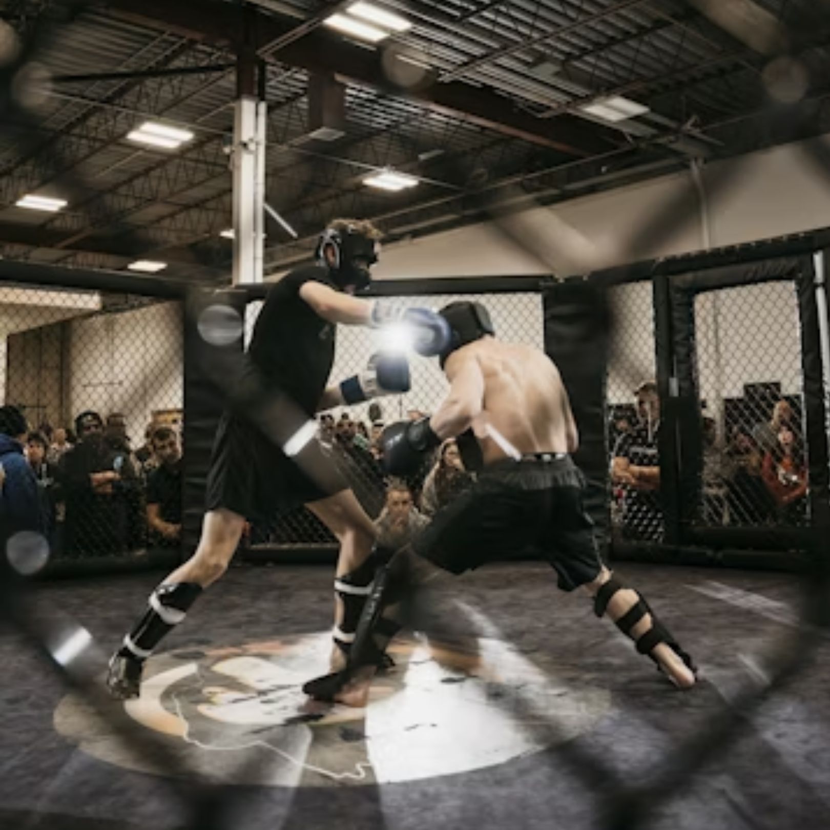 Two combatants spar in a matted MMA cage surrounded by a chain-link fence, wearing protective gear and gloves.