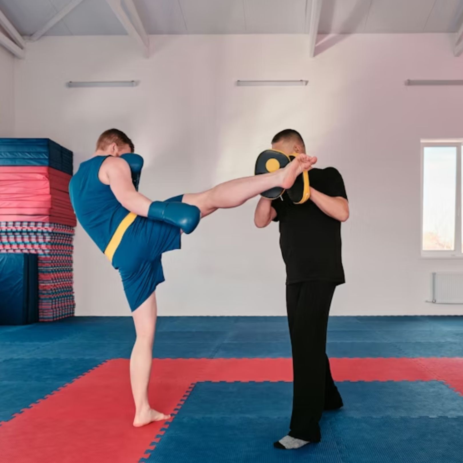 A person in a blue martial arts uniform performs a high side kick against a training pad held by an instructor.