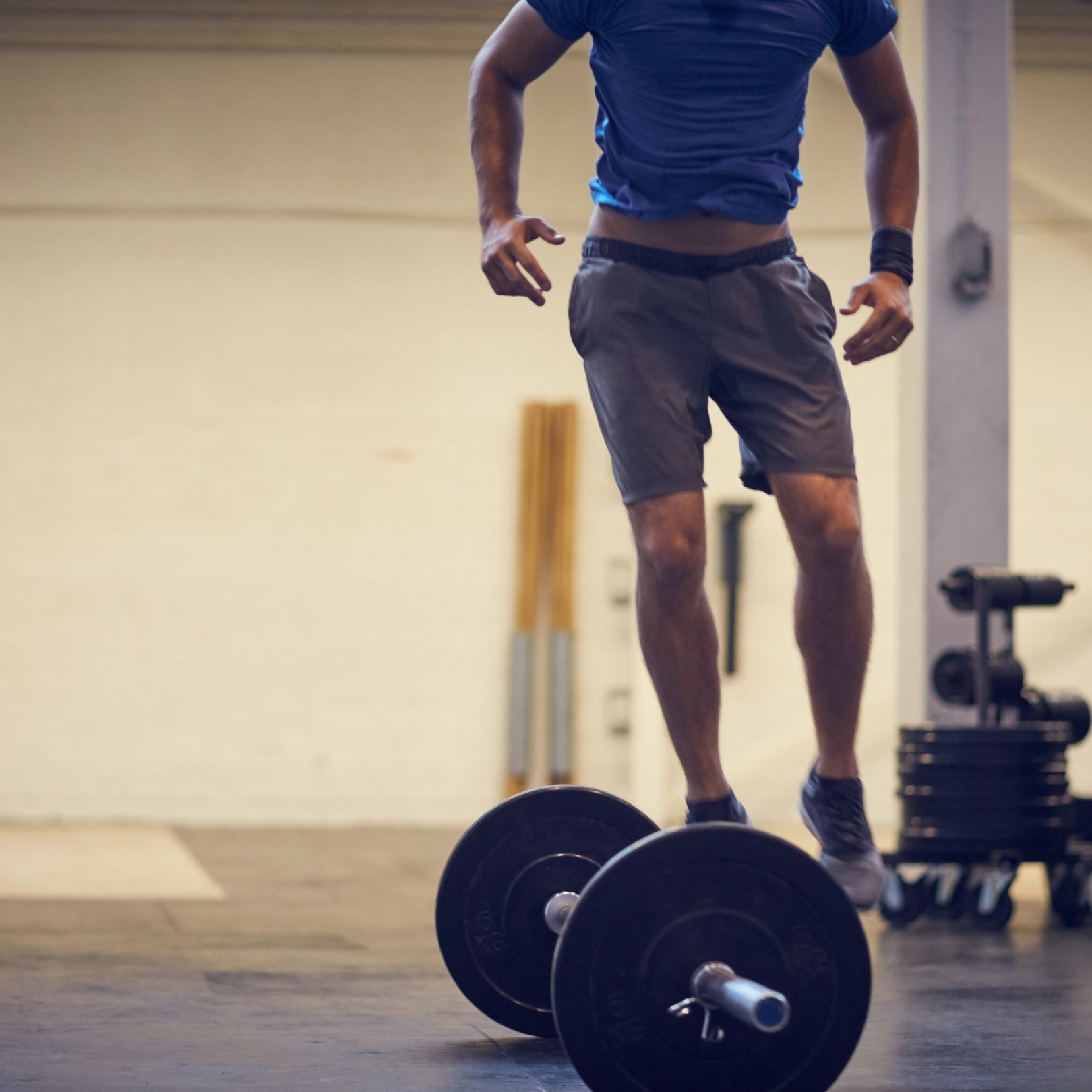 A person jumps over a barbell on the floor in a gym, wearing a blue shirt and grey athletic shorts.