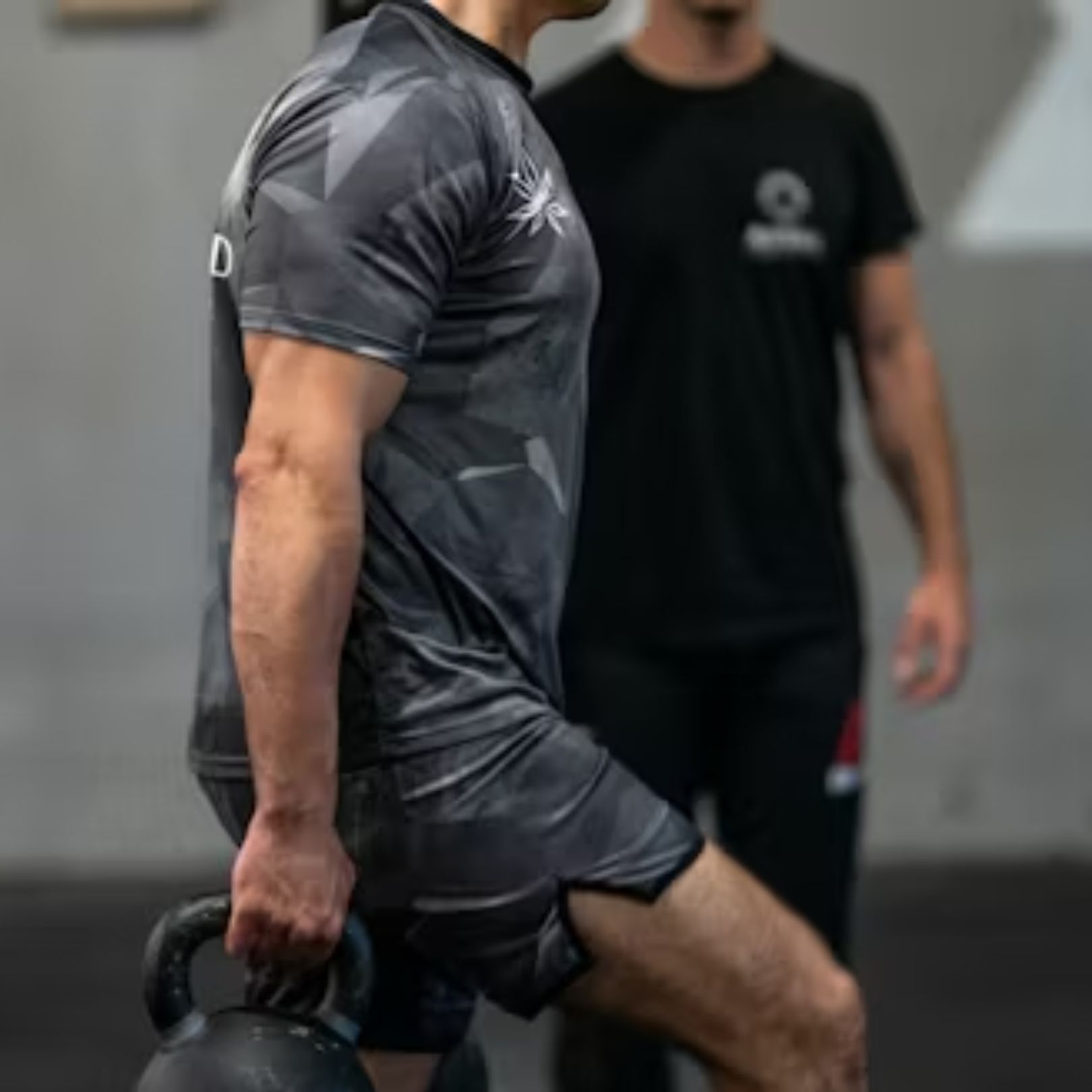 A person in a gray patterned athletic outfit performs a kettlebell lunge in a gym, watched by an instructor.