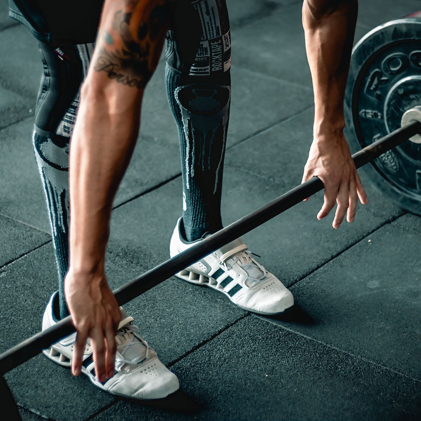A person in patterned leggings and white sneakers preparing to lift a barbell in a gym.