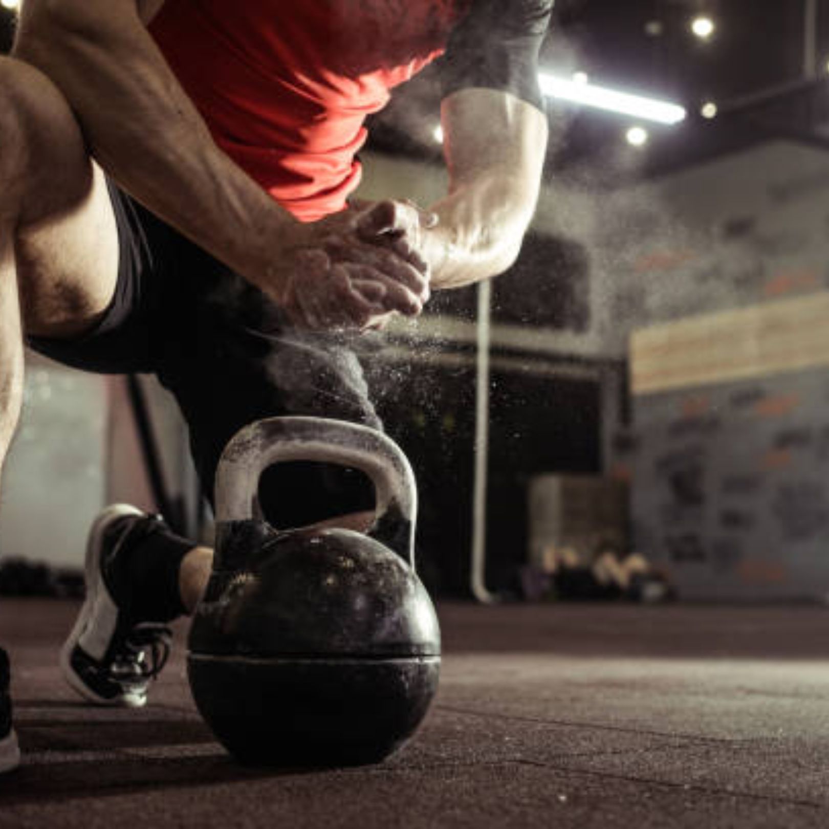 A person in a red shirt claps their hands together in a gym, releasing a cloud of chalk dust over a black kettlebell.