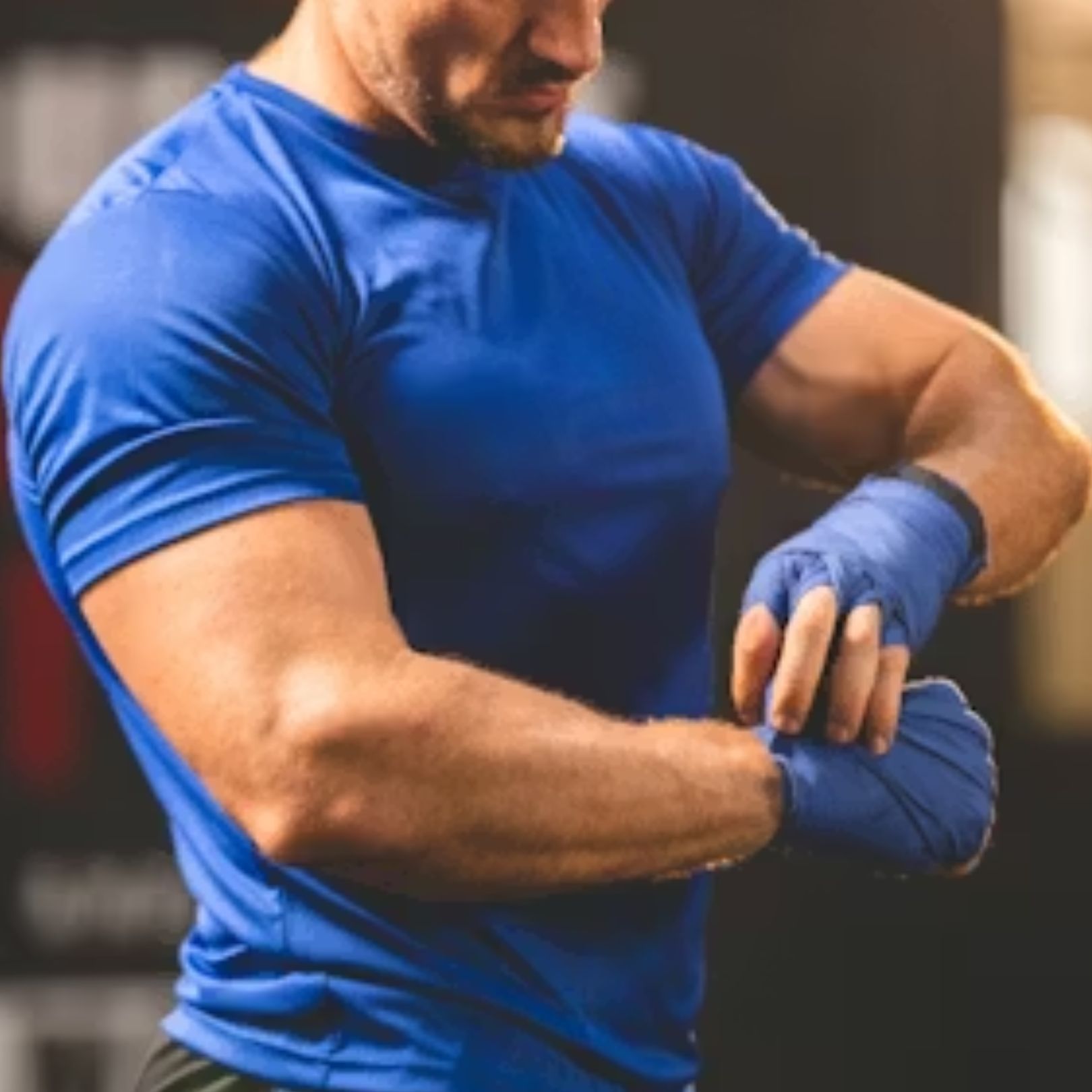 A muscular person in a blue athletic shirt wraps blue hand wraps around their wrists in a gym setting.