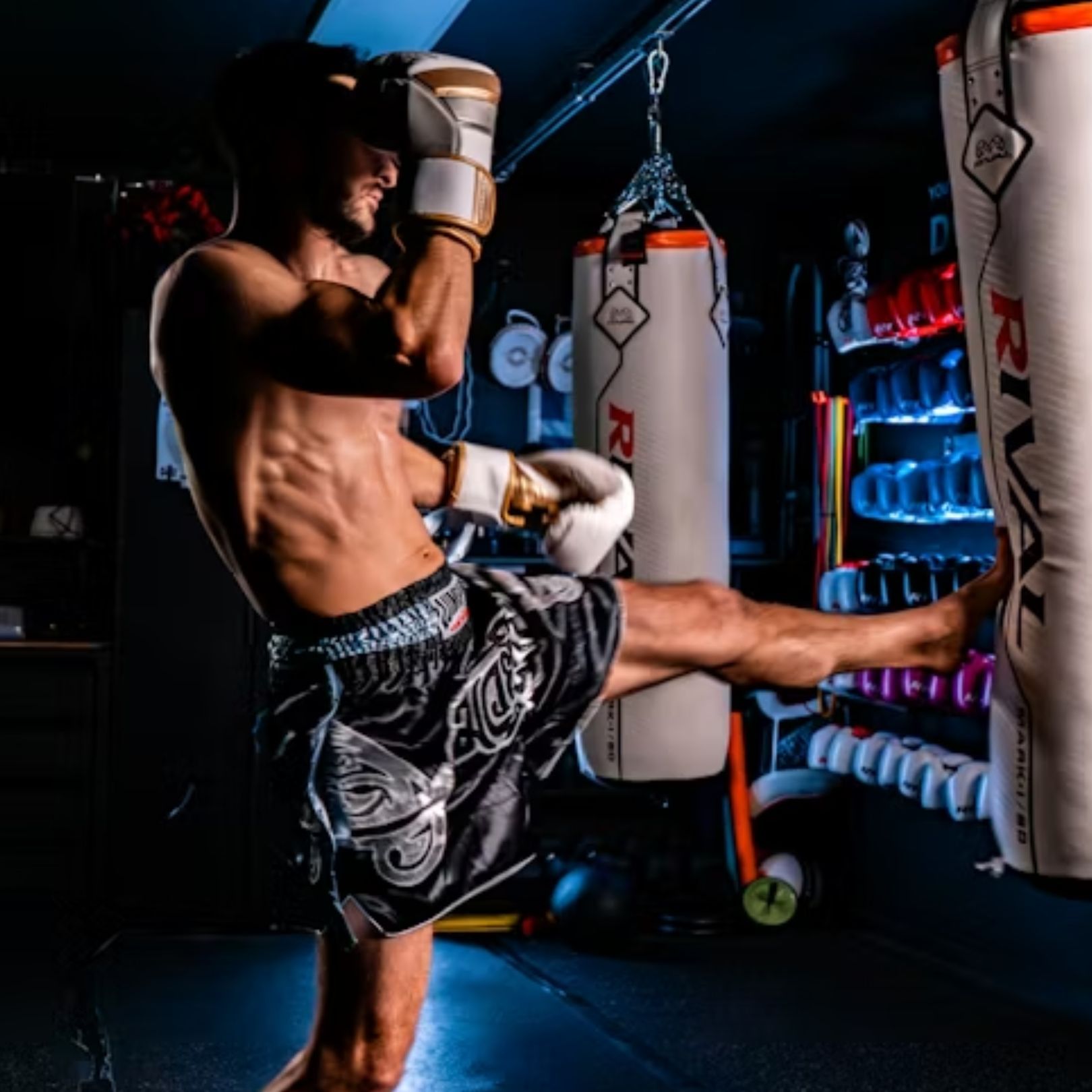 A shirtless athlete in dark Muay Thai shorts performs a side kick against a Rival punching bag in a dimly lit gym.