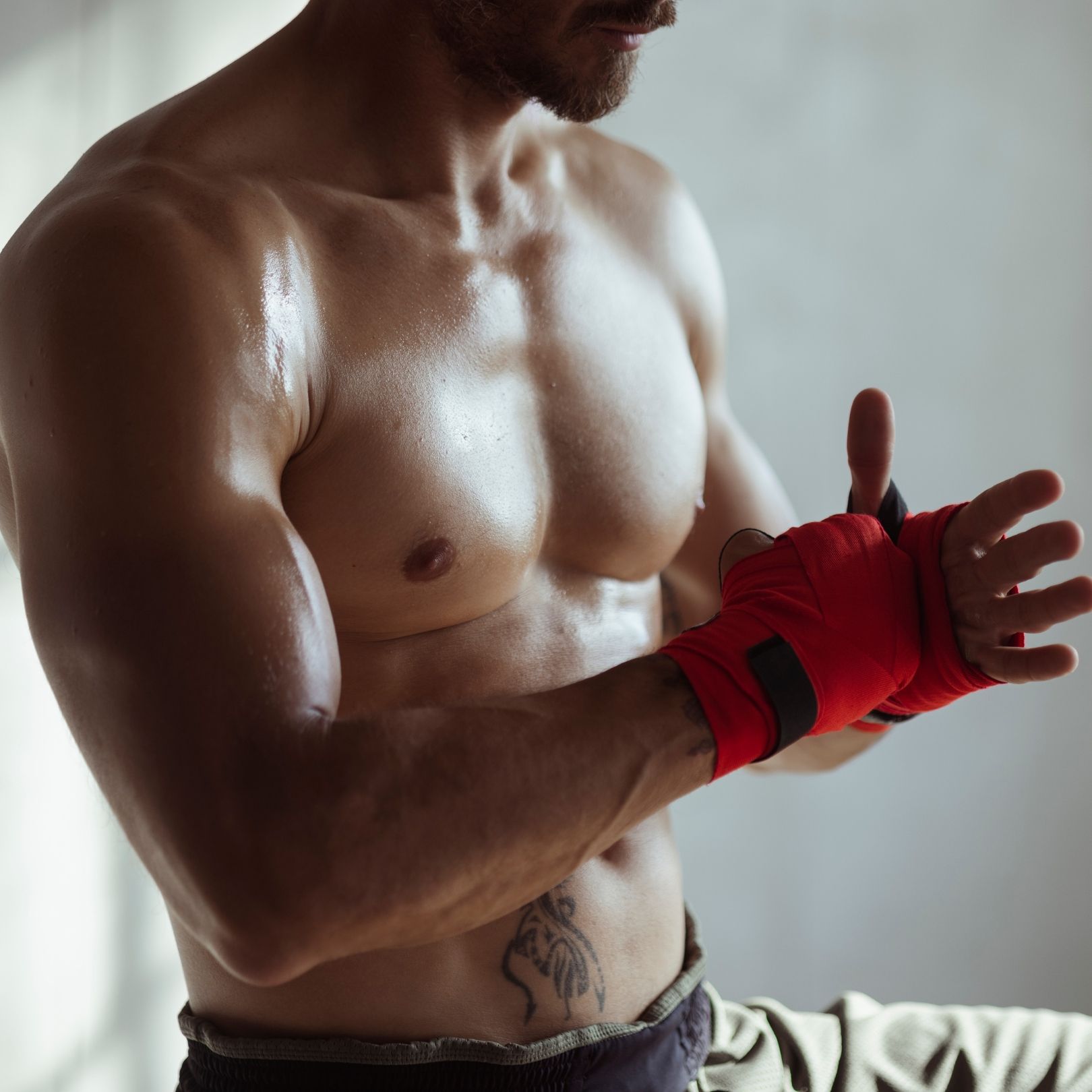 A shirtless person with a chest tattoo wearing red hand wraps, adjusting them in preparation for a workout.