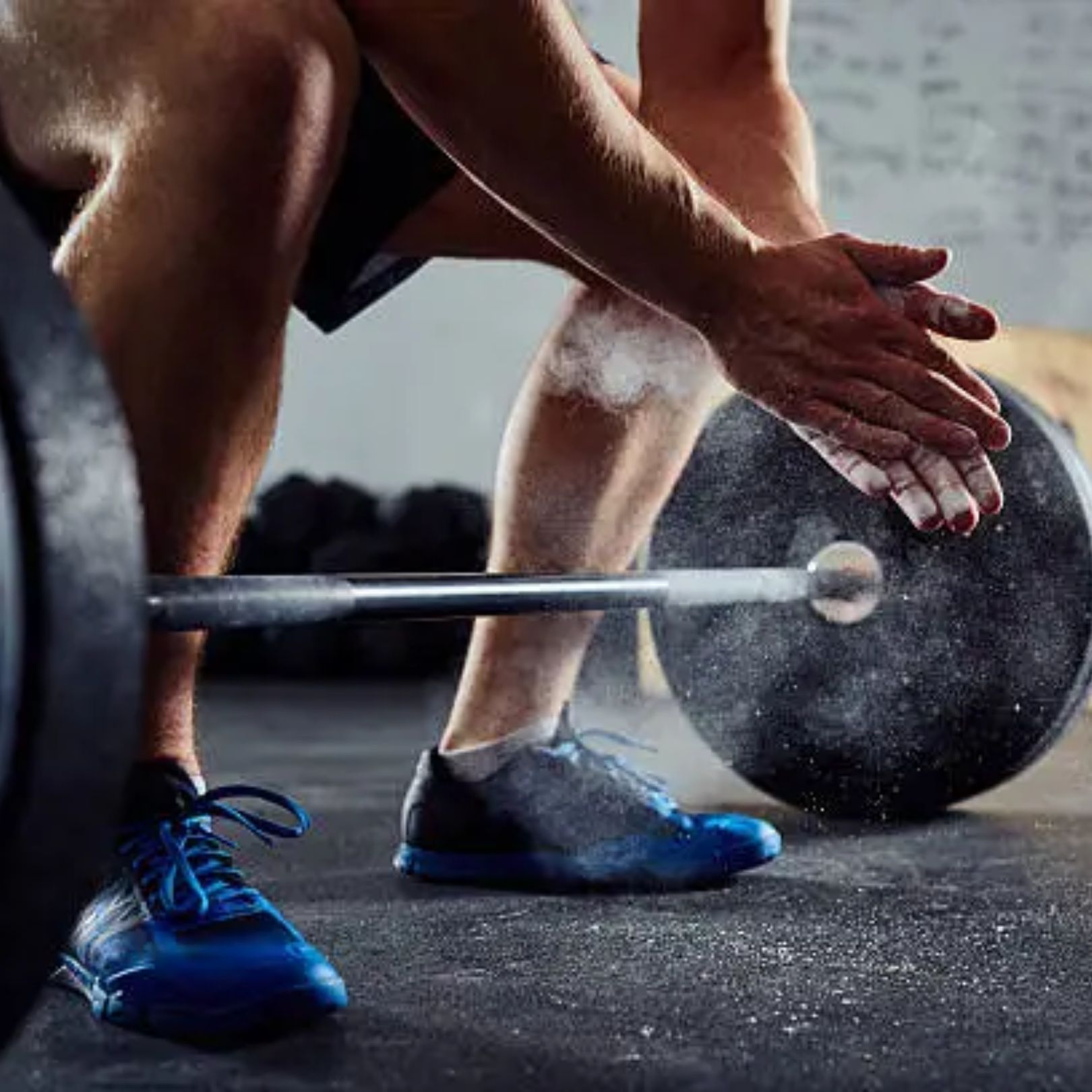 A person sits in a gym, clapping their hands together and creating a cloud of chalk dust before lifting a heavy barbell.