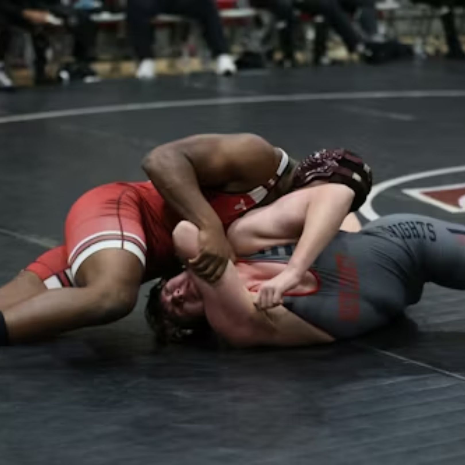 Two wrestlers in red and gray singlets grappling on a black mat during a competitive match.