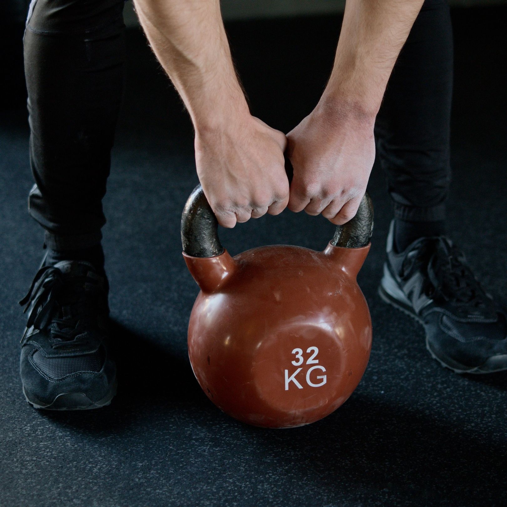 A person wearing black athletic gear grips a 32 kg kettlebell on a dark gym floor.