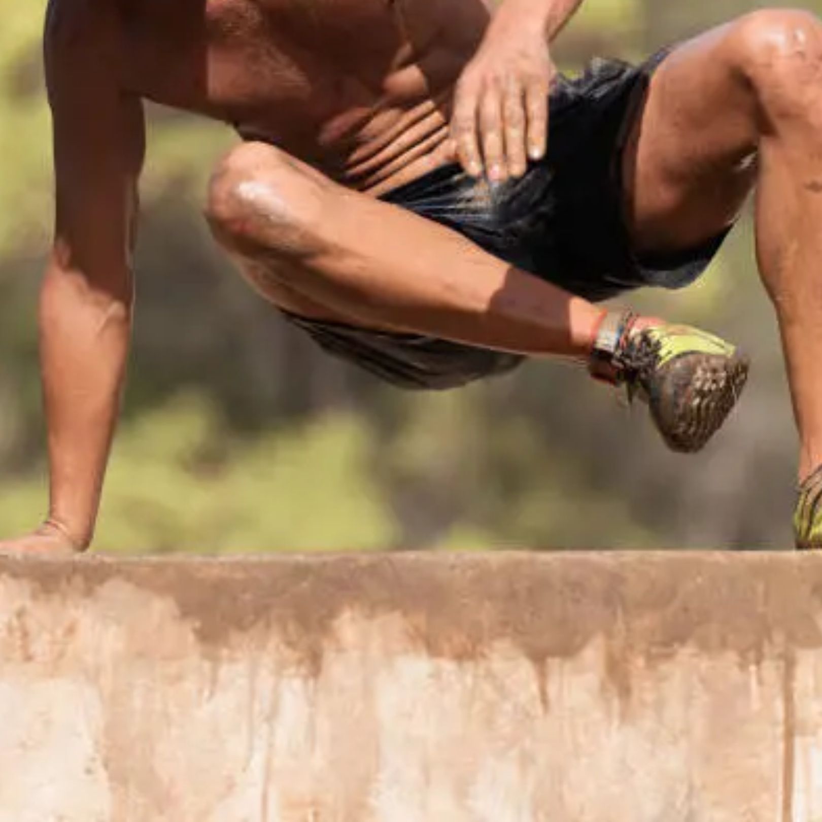 An athlete leaps over a wall, torso bent with arms and legs tucked, wearing black shorts and neon green running shoes.