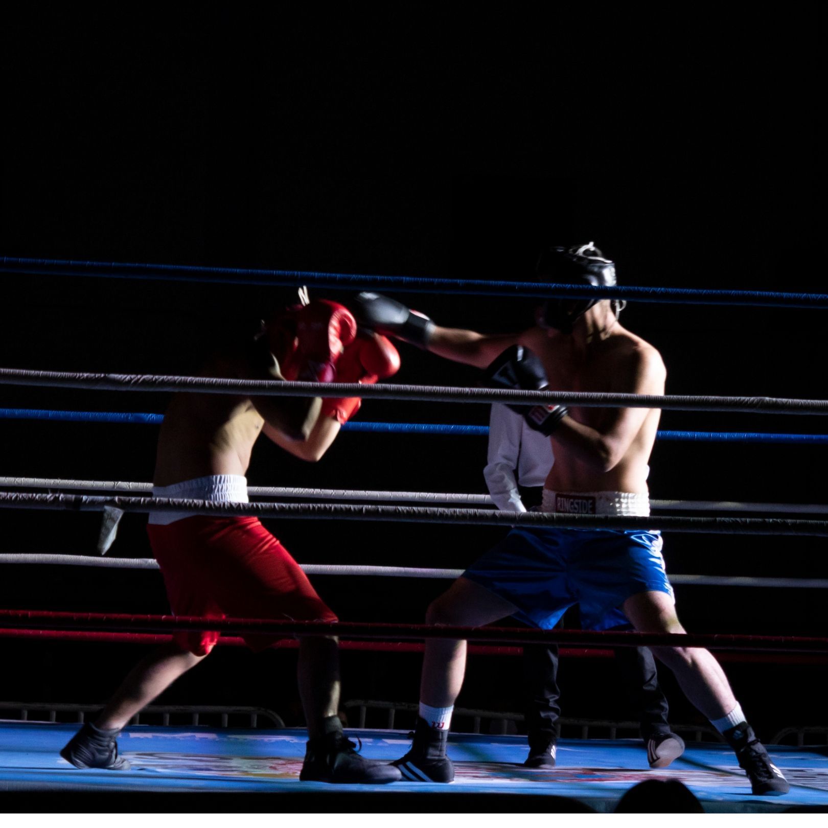 Two boxers in a ring, one wearing red shorts and the other blue, sparring under spotlights.
