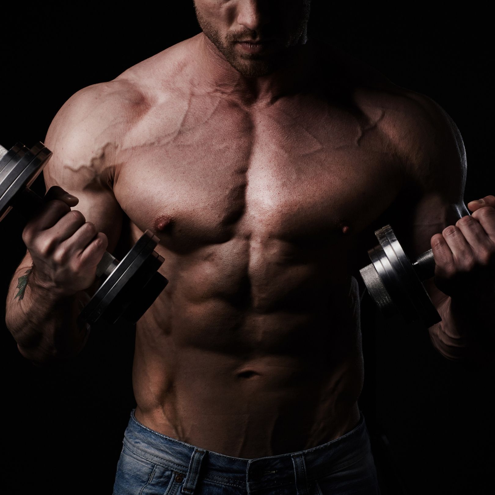 A muscular person in blue jeans performs a dumbbell curl against a dark background.