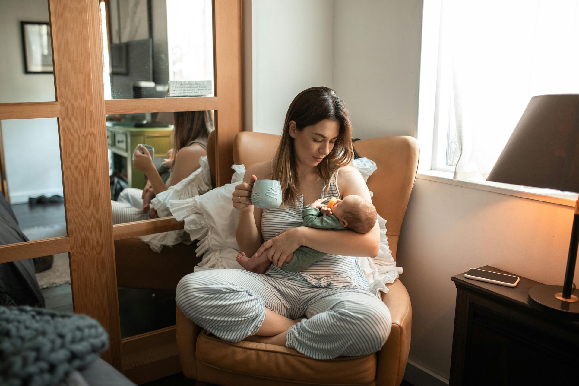 A mom holding her baby while enjoying a cup of tea that her postpartum doula made for her