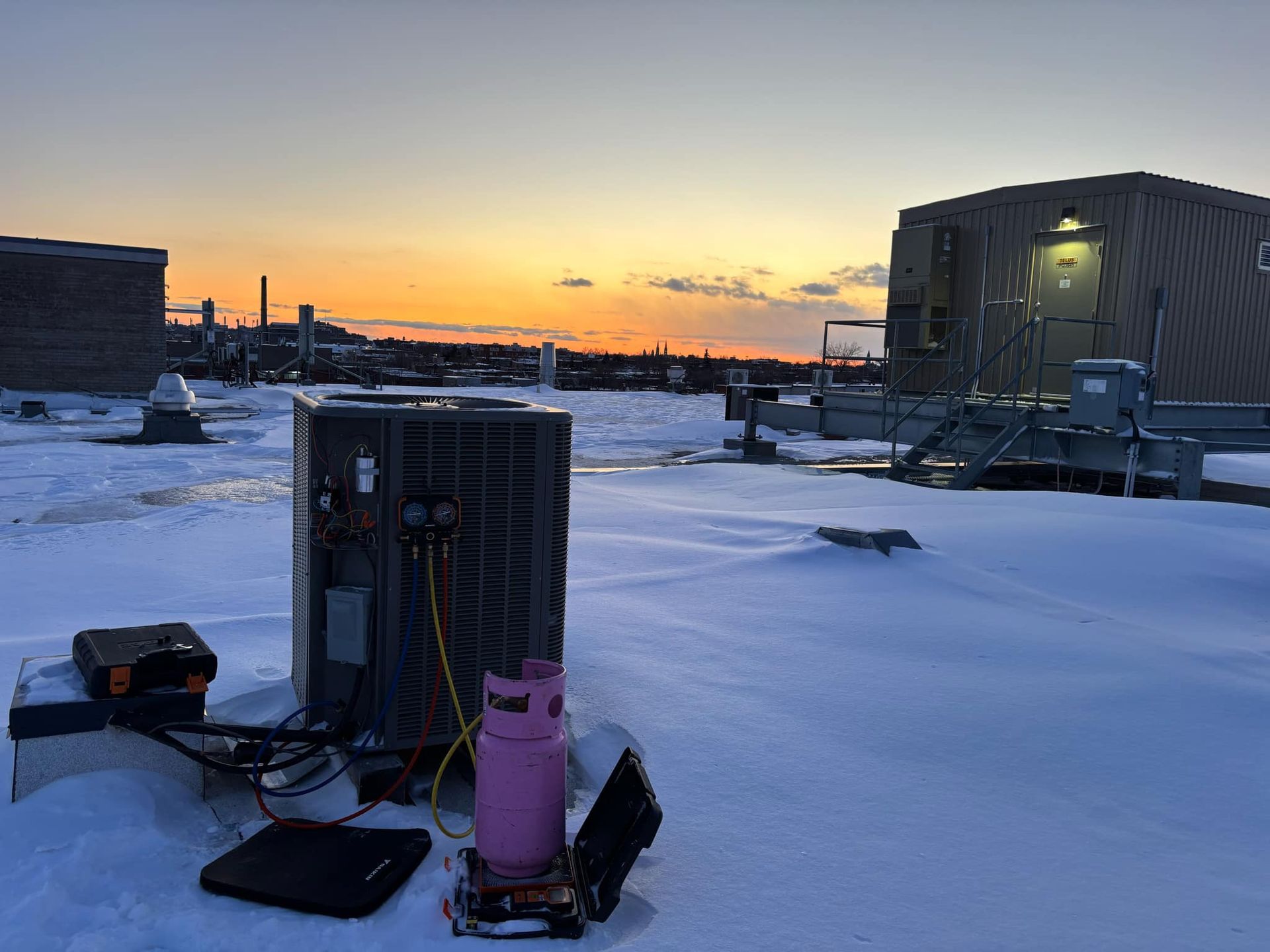 Unité de climatisation sur un toit enneigé au coucher du soleil, avec l'équipement d'un technicien.