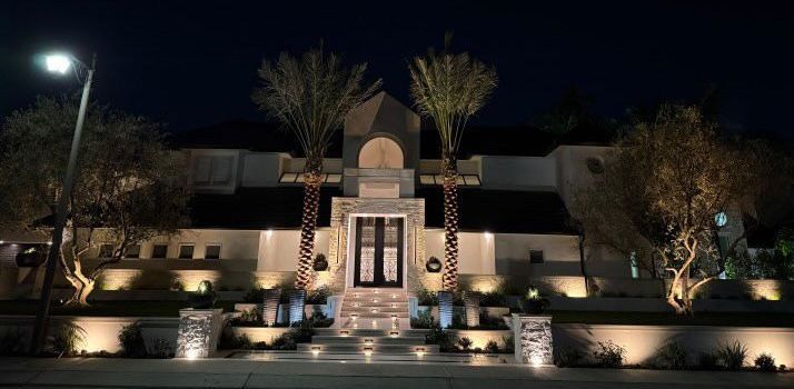 A large house is lit up at night with palm trees in front of it.