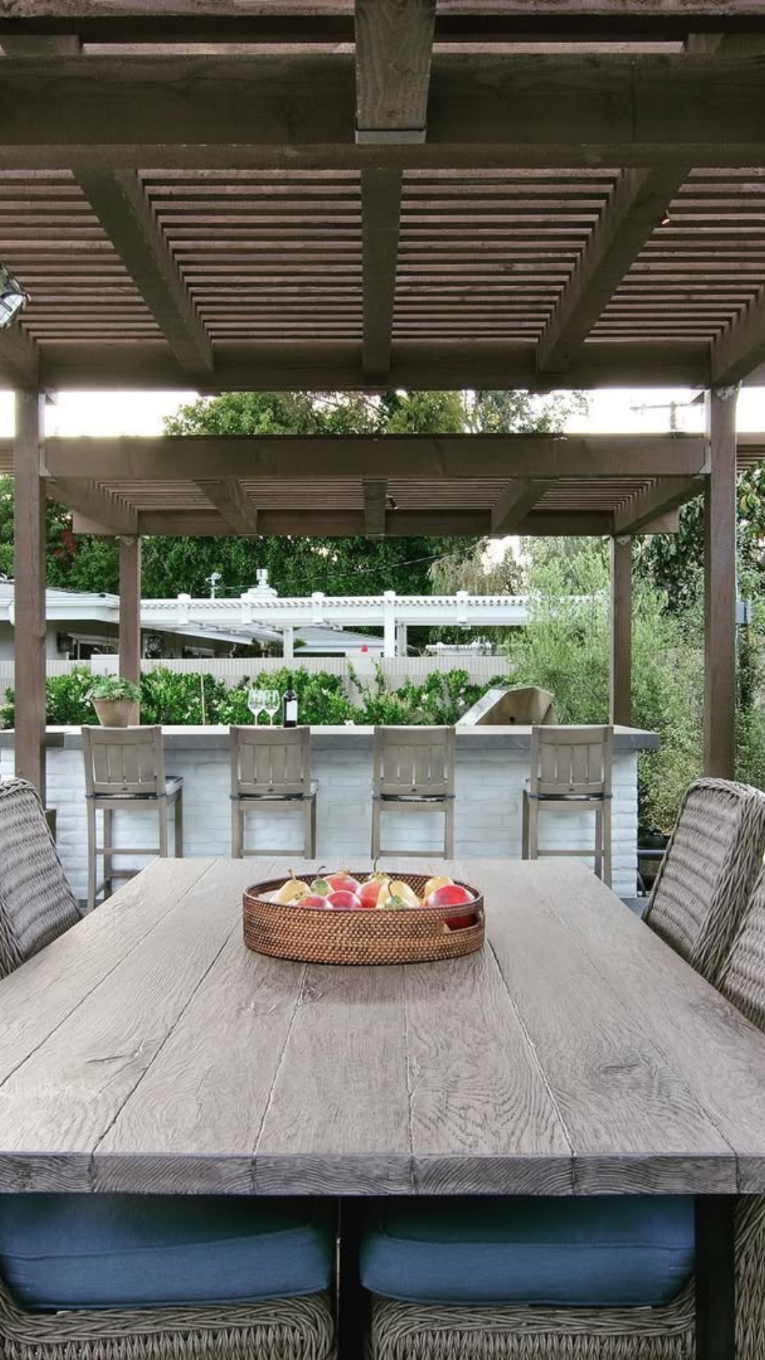 A wooden table with a tray of fruit on it under a pergola.