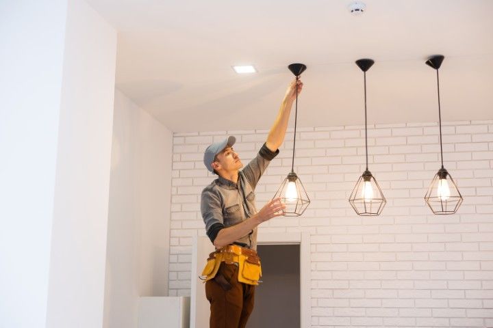 A technician standing on a ladder adjusts a pendant light fixture in a modern room with white brick walls.