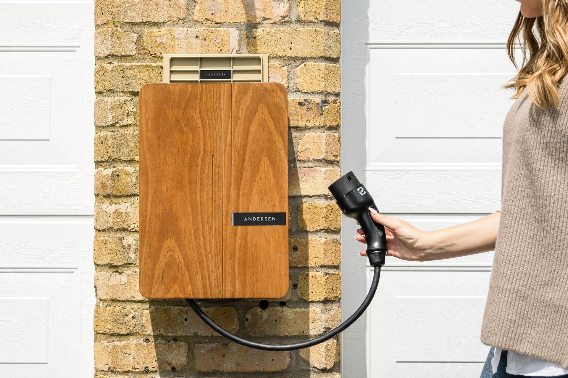 A person holds the charging cable of a wood-paneled electric vehicle home charger mounted on a brick wall.