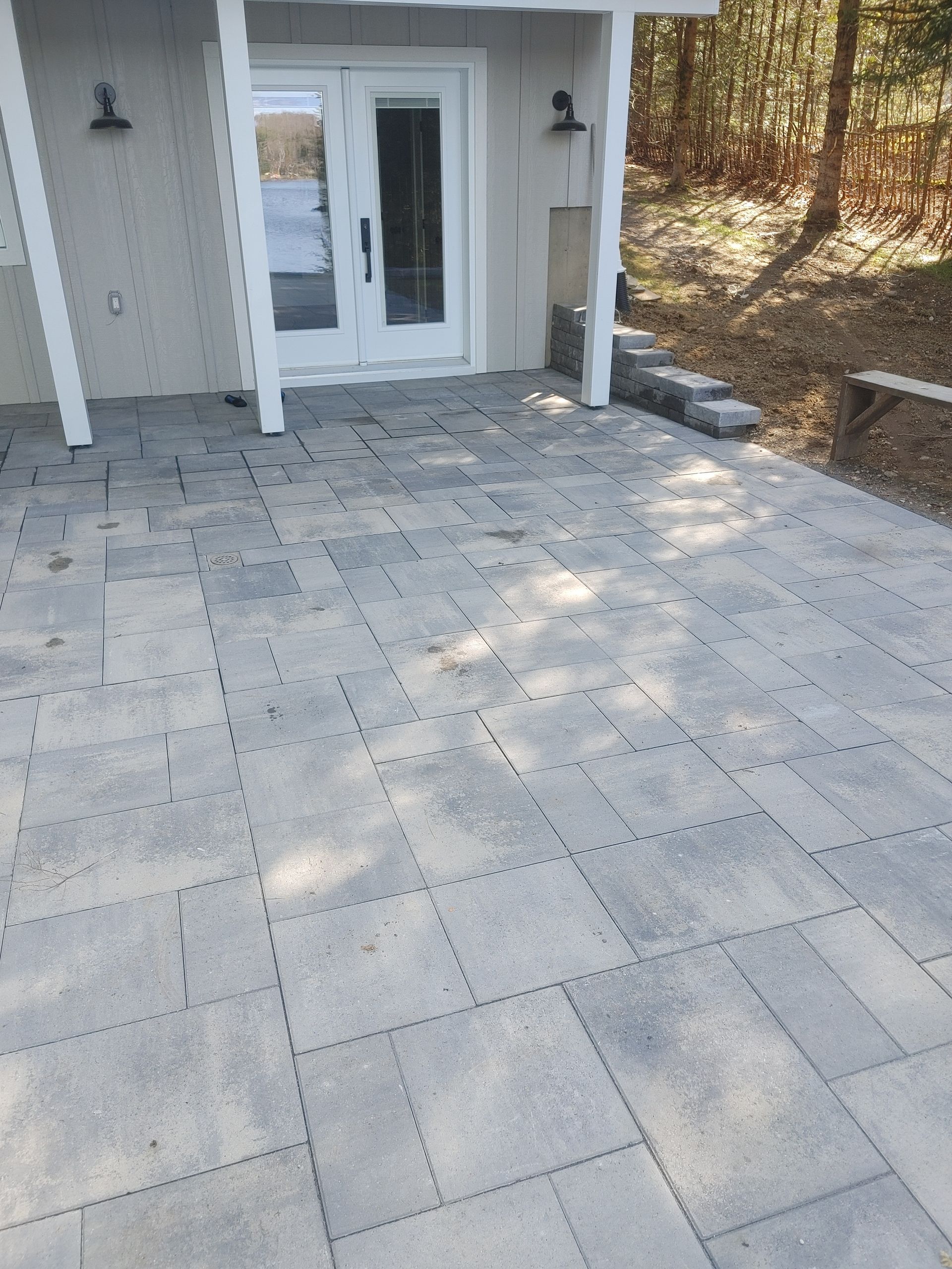 Terrasse en pierre grise devant un bâtiment aux portes blanches ; forêt en arrière-plan.
