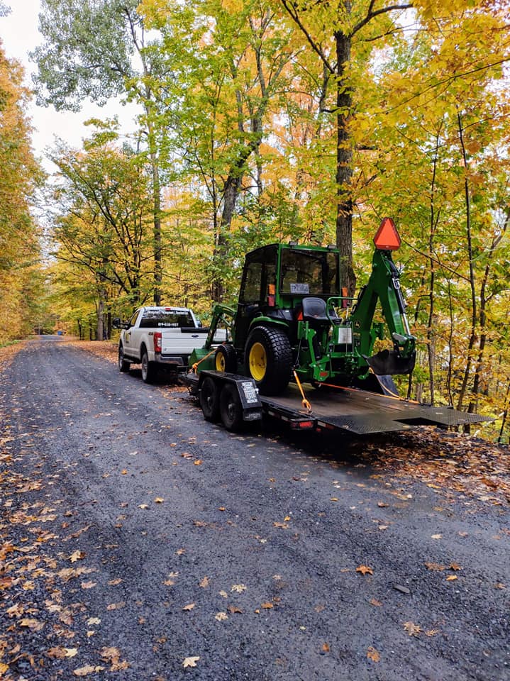 Un camion blanc tractant une remorque avec un tracteur John Deere vert circule sur un chemin de gravier.