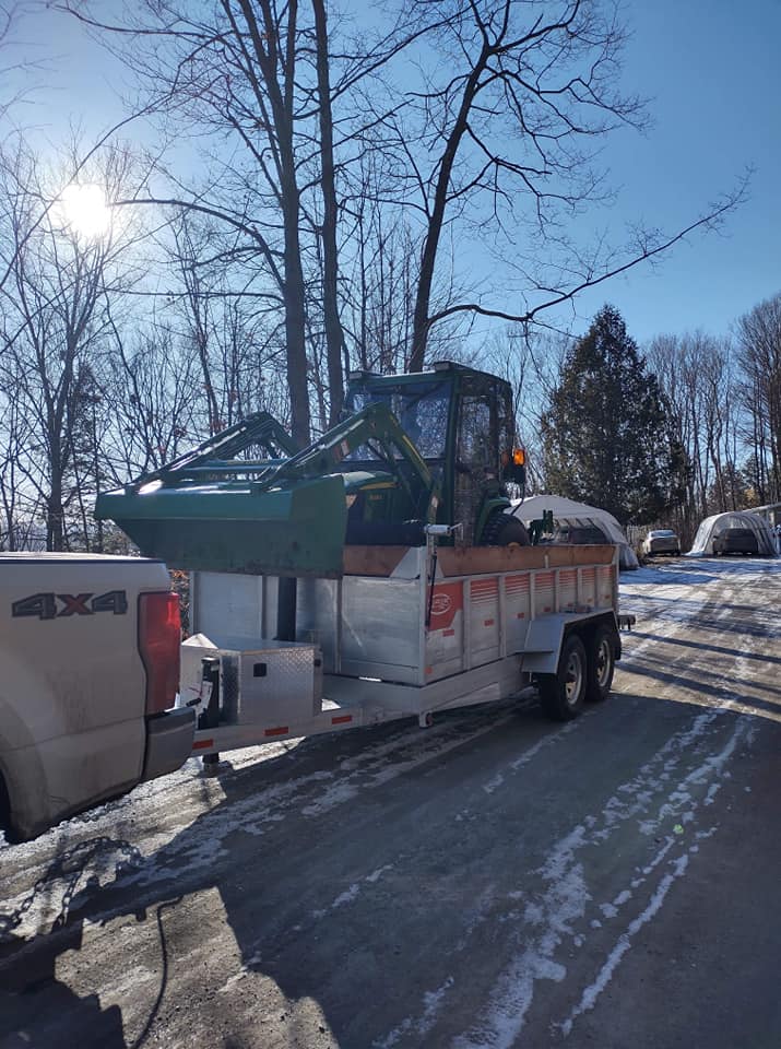 Un tracteur équipé d'un chargeur frontal est attelé à une remorque elle-même attelée à un camion, sur une route enneigée.
