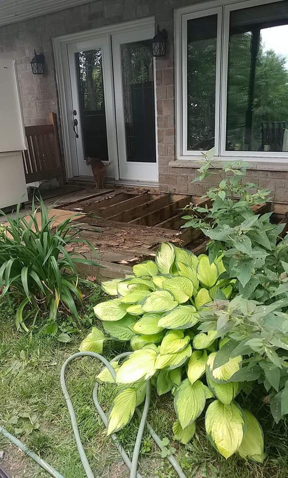 Porche en bois effondré devant une maison. Trou dans la terrasse, avec de la verdure au premier plan.