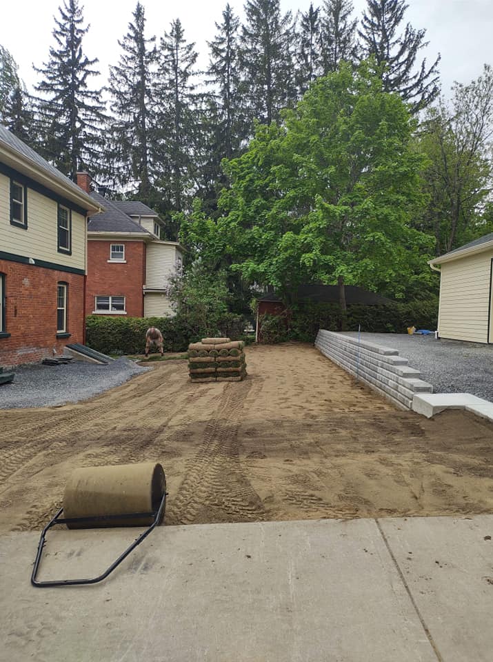 Préparation du gazon, rouleaux de gazon prêts à être posés. Un rouleau est posé au sol ; bâtiments et arbres en arrière-plan.