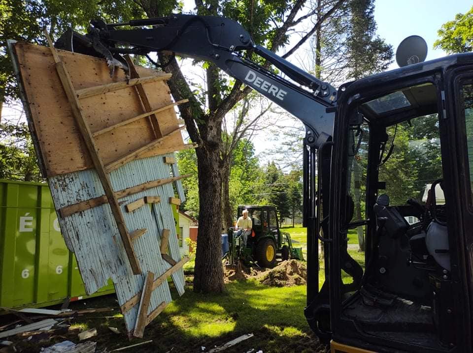 Une pelle mécanique John Deere soulève une partie d'une structure en ruine. Des hommes travaillent à l'arrière-plan. 