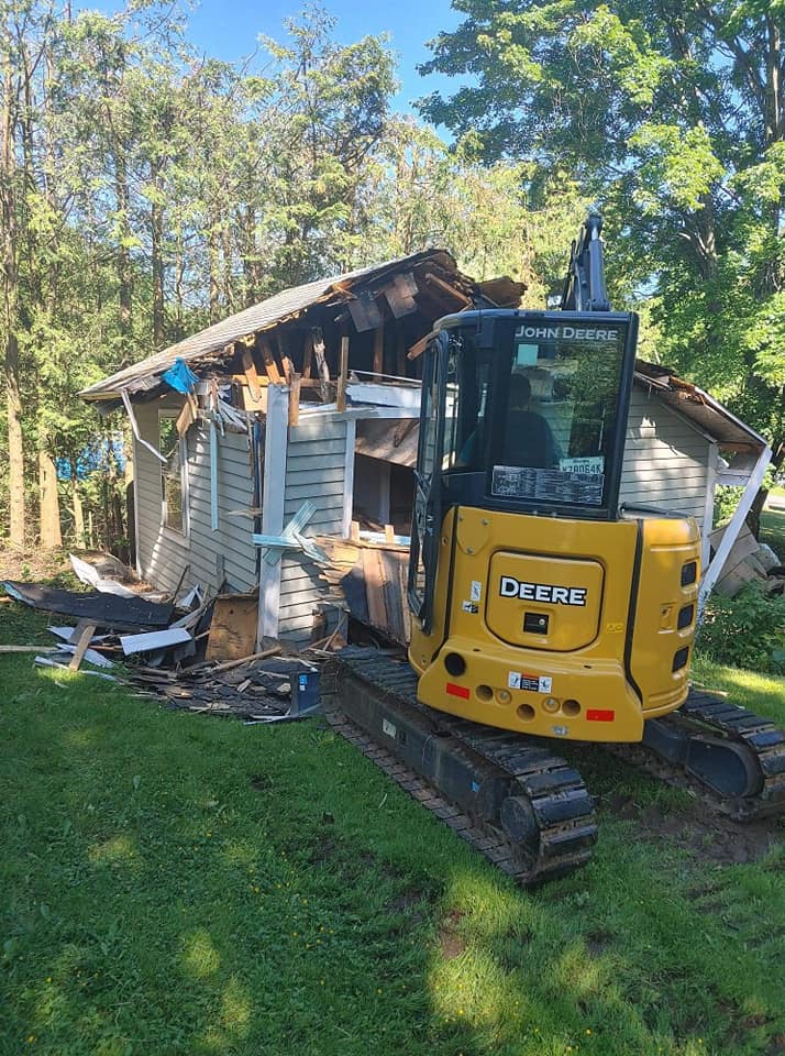 Une pelleteuse jaune démolit une petite maison sur une pelouse avec des arbres en arrière-plan.