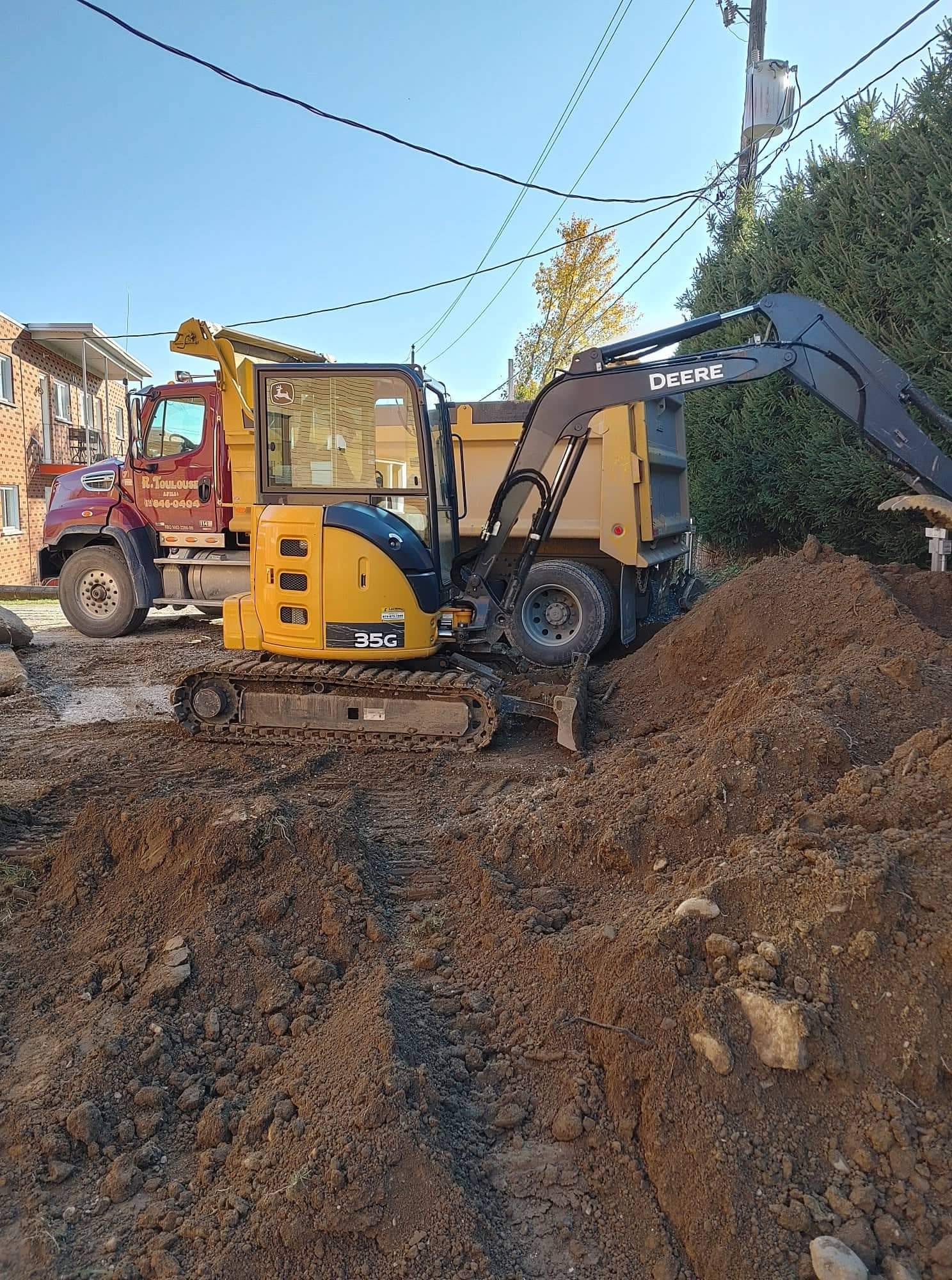 Une pelleteuse jaune creuse la terre ; un camion rouge est visible en arrière-plan.