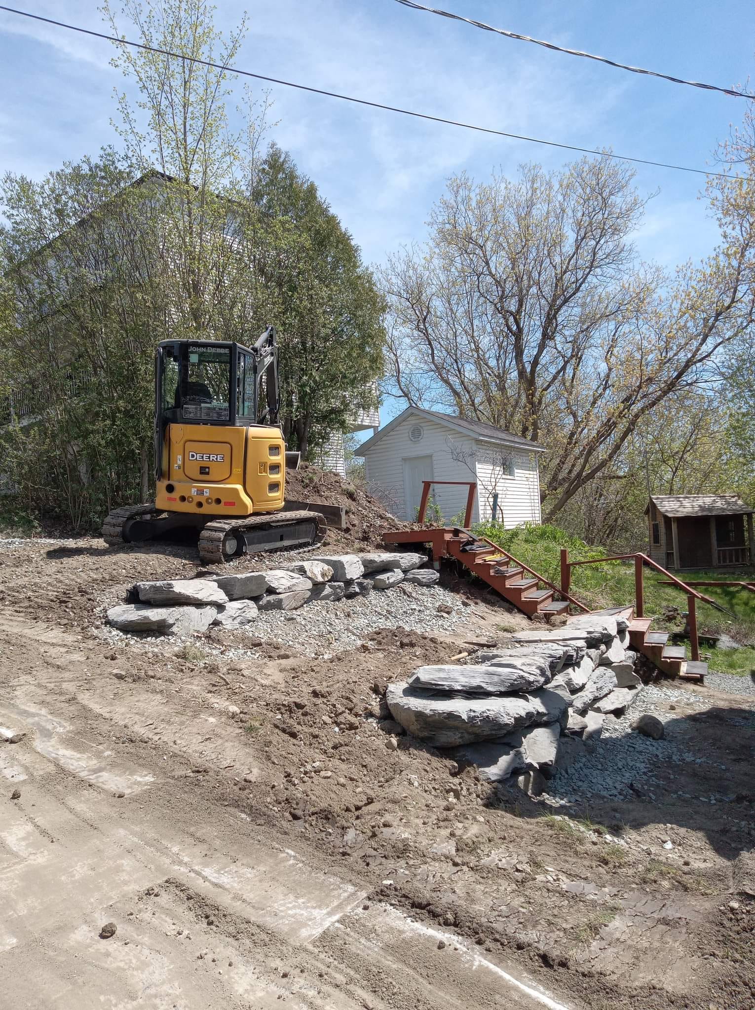Mini-pelle sur un talus de terre, construisant un escalier en pierre et en brique, avec un bâtiment blanc en arrière-plan.