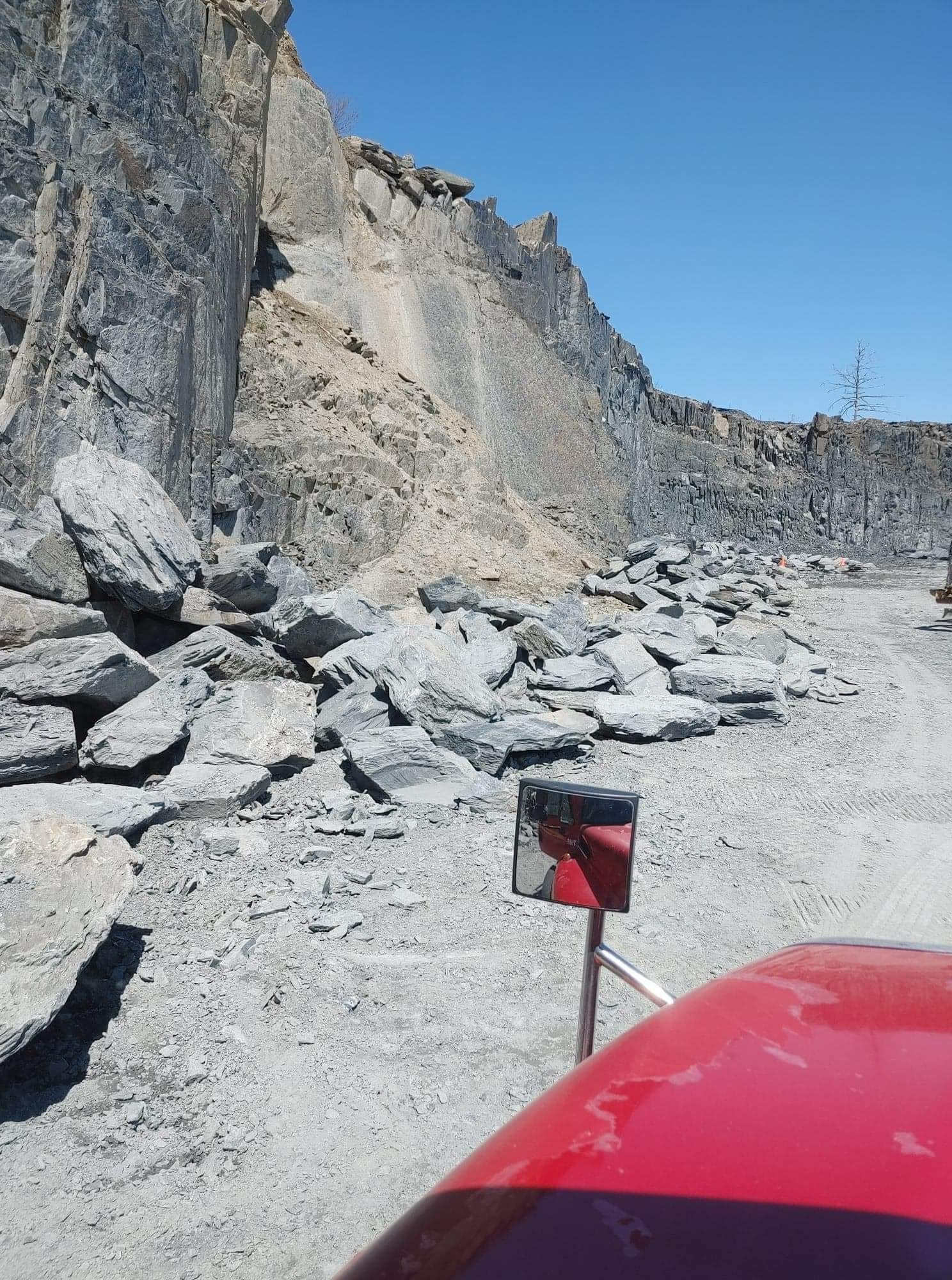 Vue depuis un véhicule rouge d'une falaise rocheuse et d'un tas de roches grises sur un chemin de gravier sous un ciel bleu.