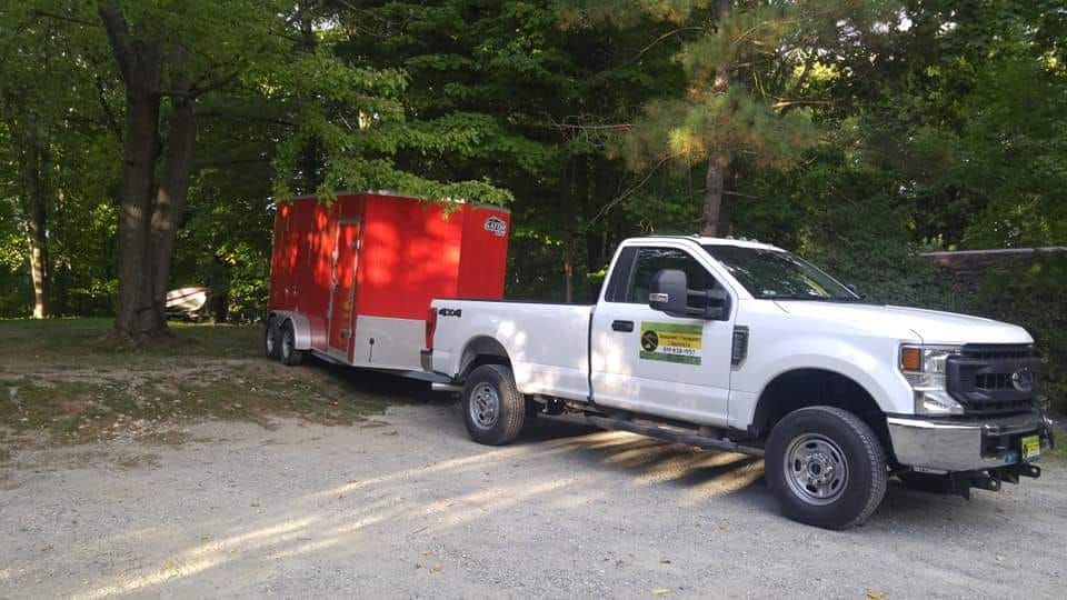 Une camionnette blanche tracte une remorque rouge sur une allée de gravier, avec des arbres en arrière-plan.
