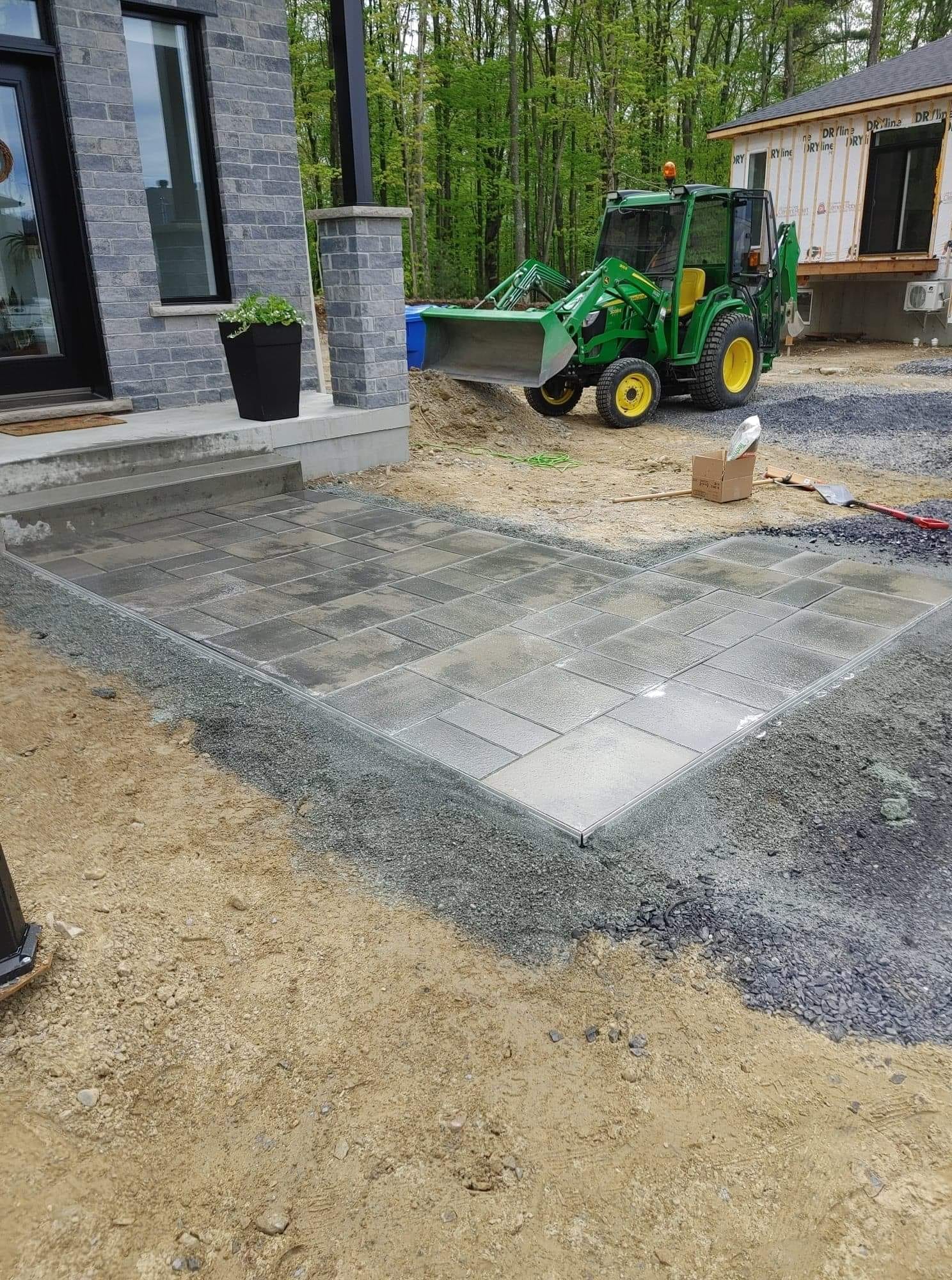 Aménagement d'une terrasse : dalles grises posées, entourées de gravier, devant une maison, petit tracteur en arrière-plan.