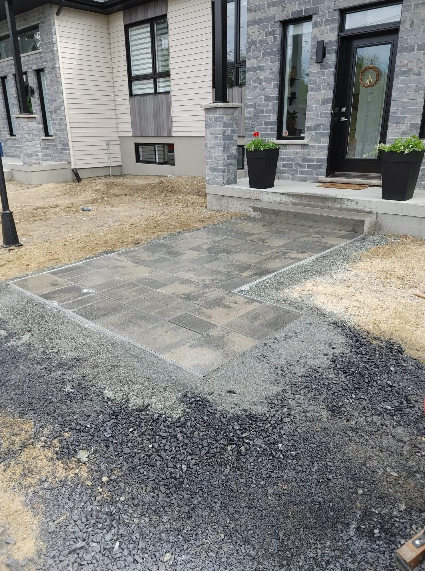 Allée nouvellement pavée menant à l'entrée d'une maison ; façade en pierre grise, porte sombre, plantes en pot.