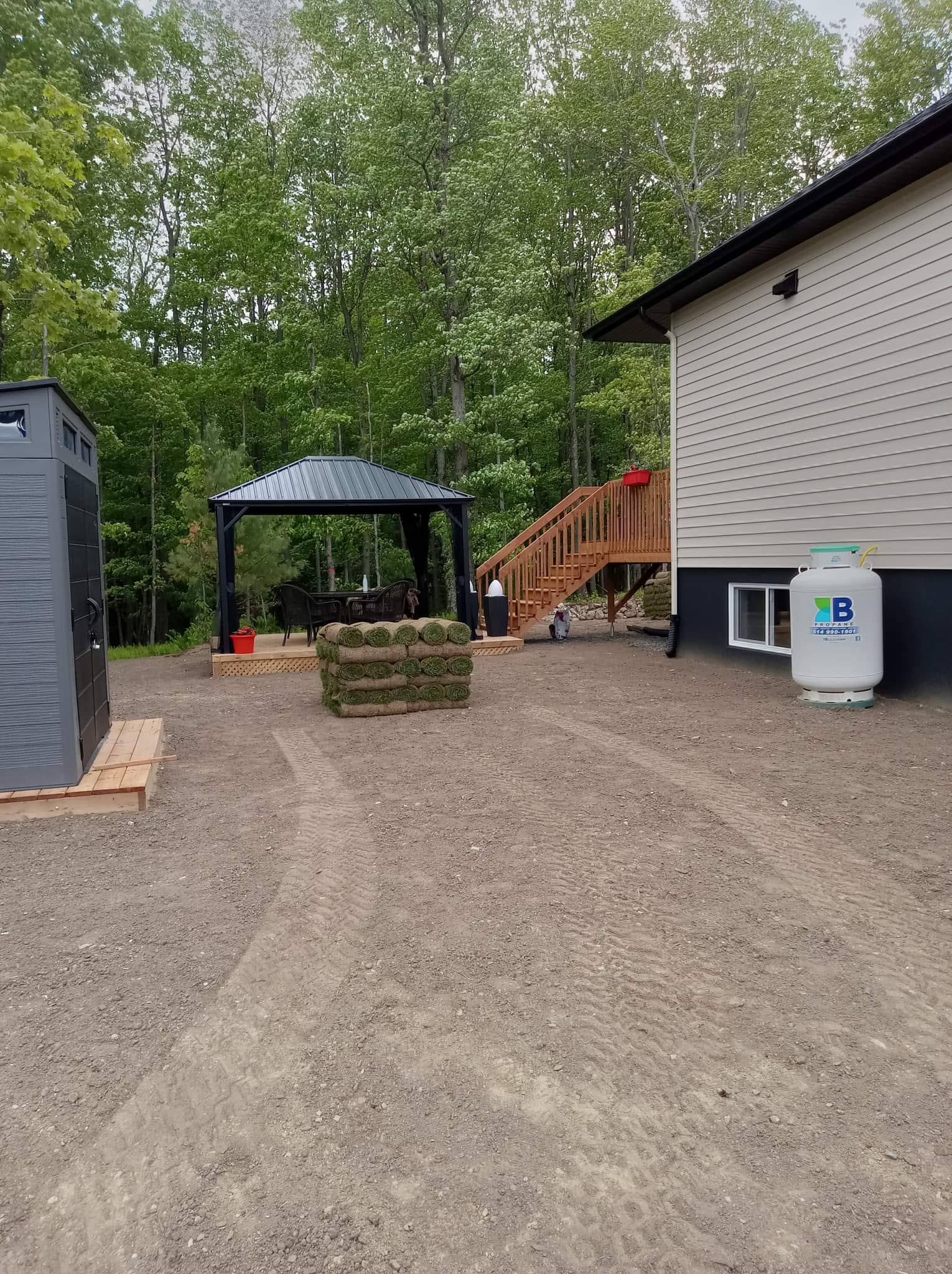 Cour en gravier avec un kiosque, une bonbonne de propane, un abri de jardin et un escalier menant à une terrasse.