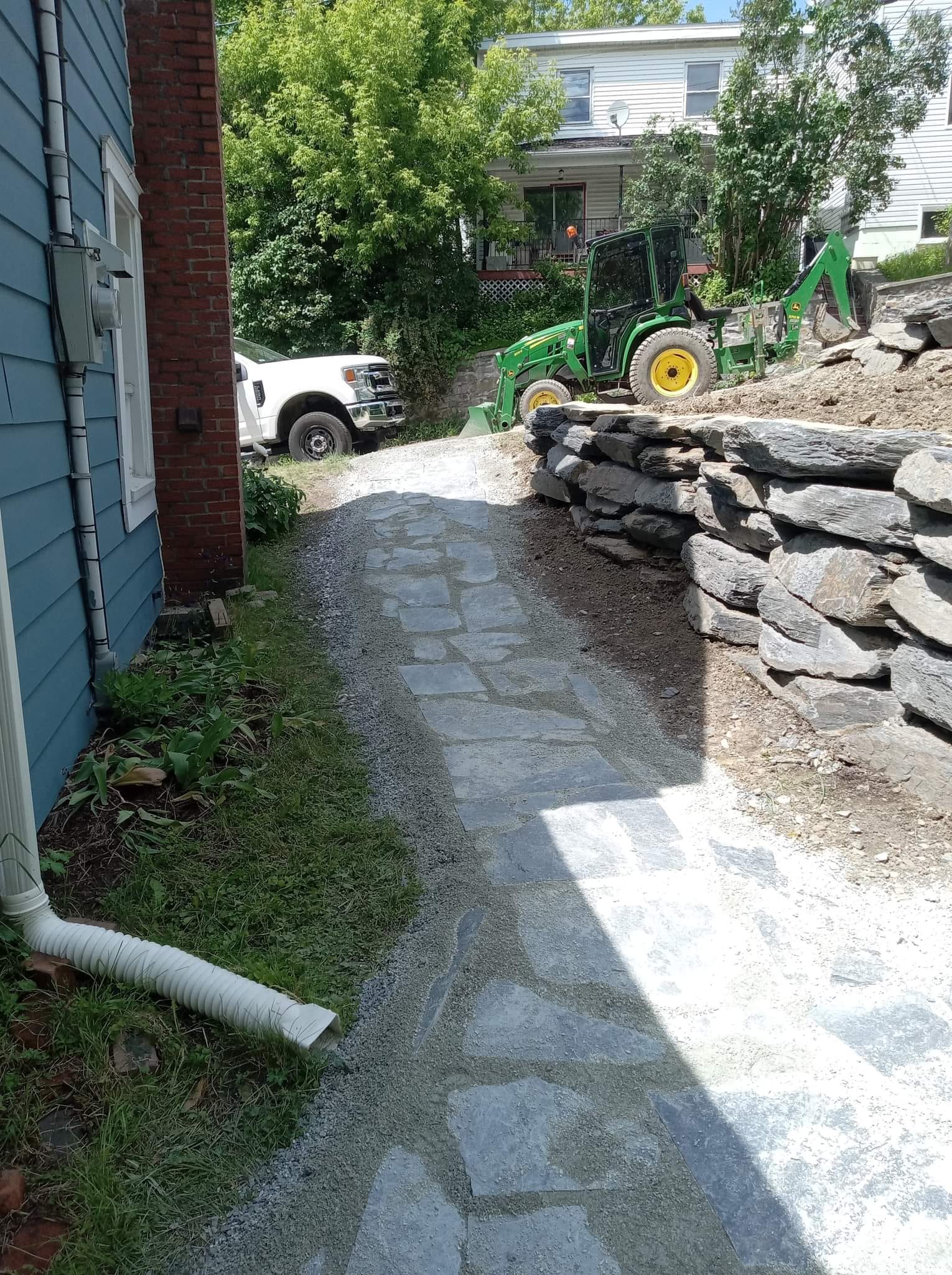 Allée de gravier jouxtant un bâtiment bleu et un mur de soutènement en pierre ; camion et tracteur visibles.