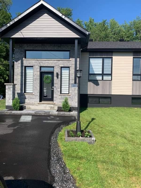 Maison moderne avec façade en briques grises, porte noire et bardage clair, avec une allée incurvée.
