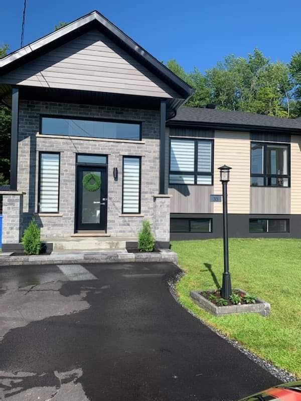 Maison moderne avec façade en briques grises, boiseries noires et allée asphaltée par une journée ensoleillée.