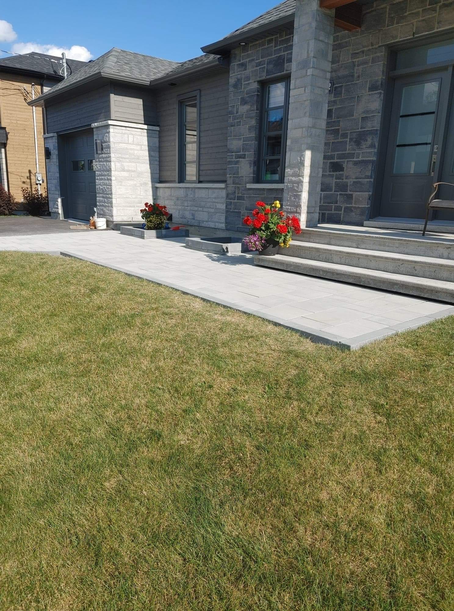 Maison en pierre grise avec allée et pelouse ; pots de fleurs rouges.