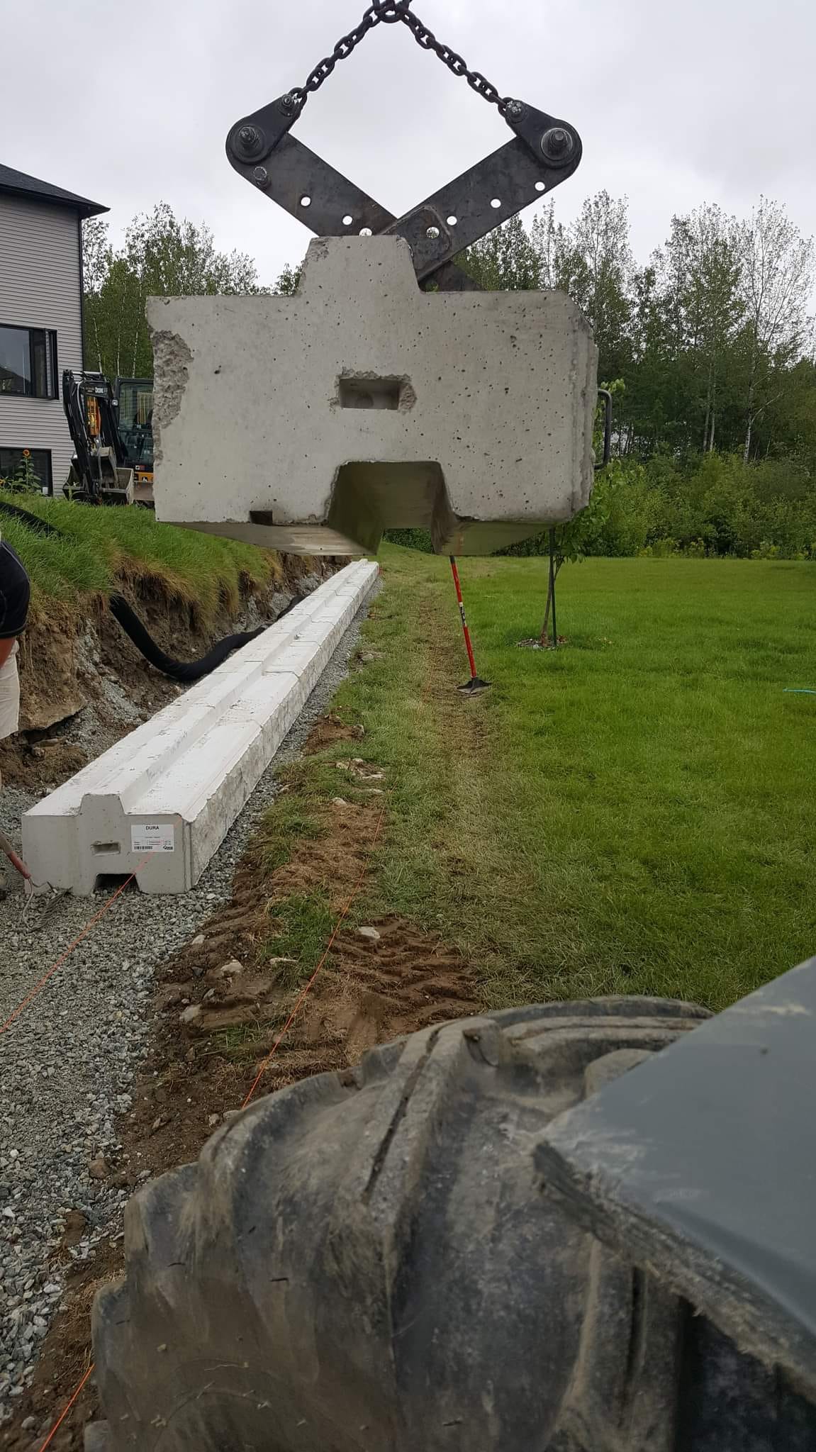 Un pan de mur en béton est soulevé par des engins lourds, à proximité d'une tranchée préparée et d'un bâtiment.