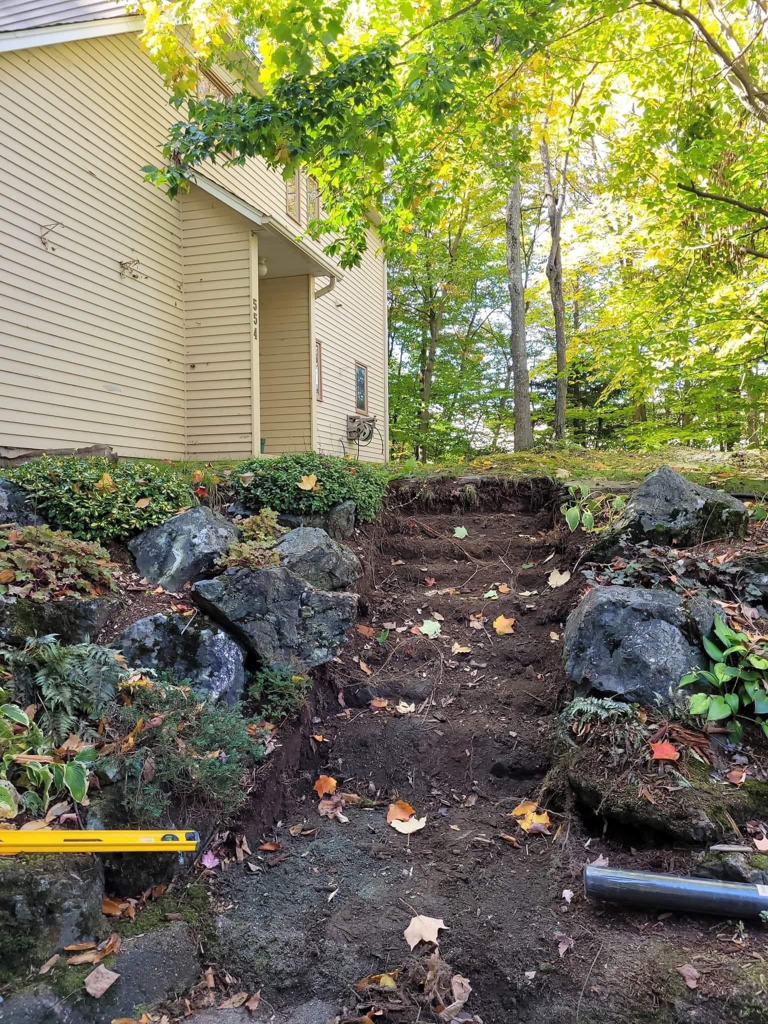 Des marches en pierre, recouvertes de terre et de cailloux, mènent à flanc de colline, à côté d'un bâtiment beige et d'arbres.