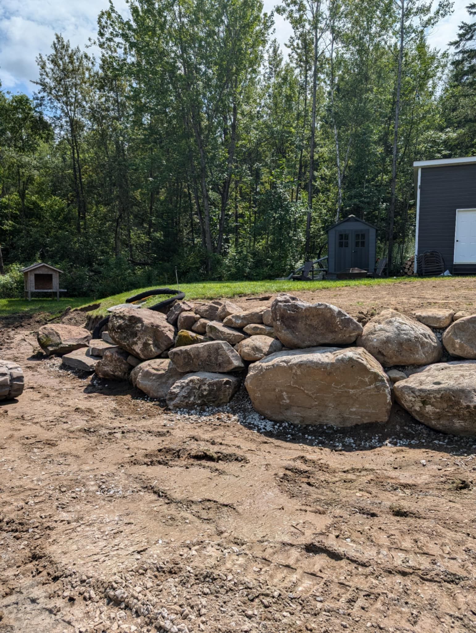 Mur de soutènement en pierre sur un talus de terre. Herbe verte et arbres en arrière-plan. Abri de jardin visible.