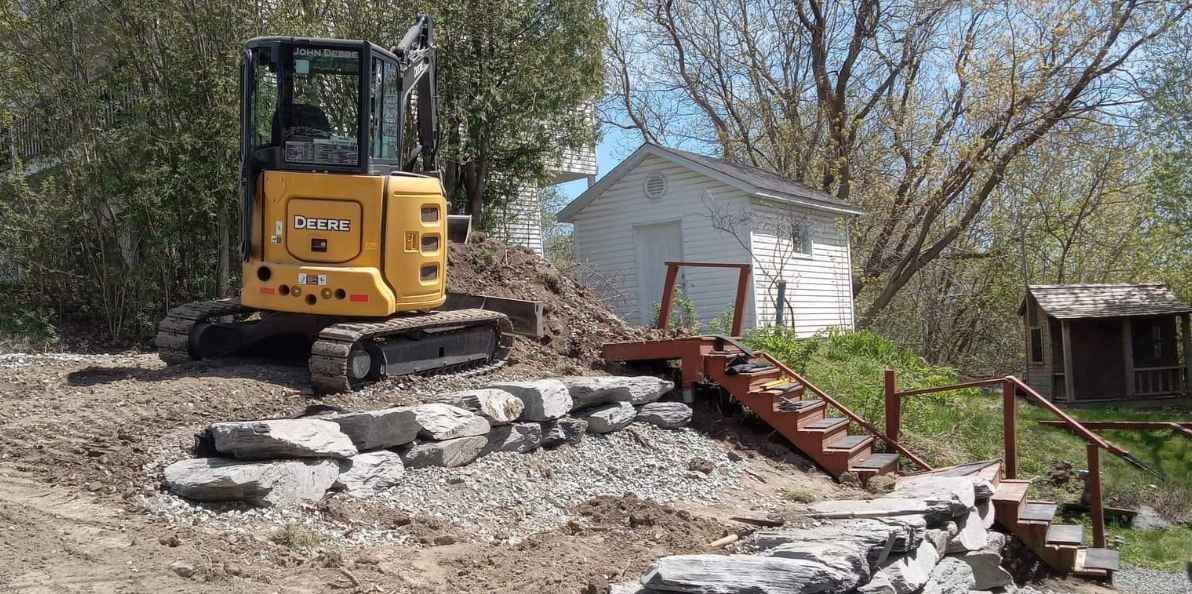 Une pelleteuse jaune se trouve sur une colline de terre près d'un mur de pierre, d'escaliers et d'un bâtiment blanc.