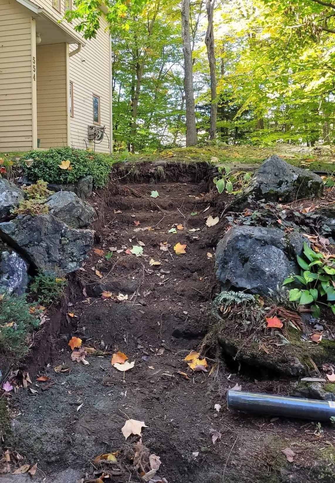 Des marches de terre taillées à flanc de colline, entourées de rochers et de végétation, près d'une maison au bardage beige.