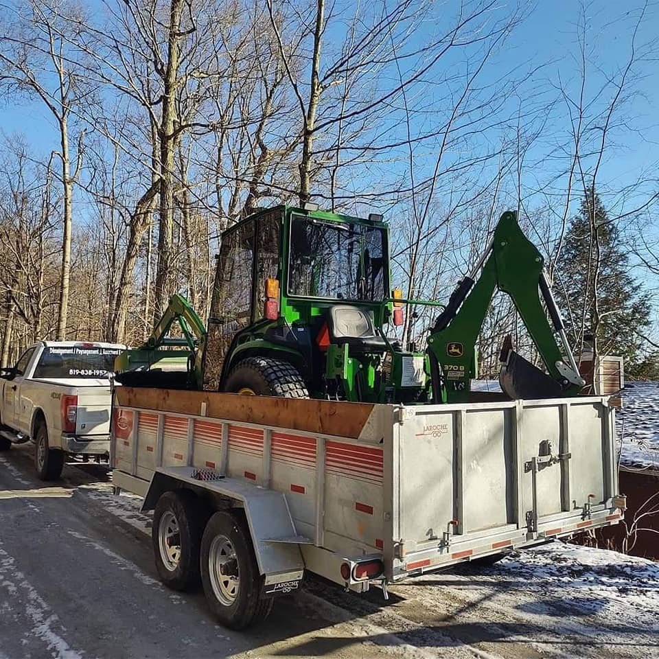 Tracteur John Deere vert sur une remorque, attelée à une camionnette blanche, sur une route.