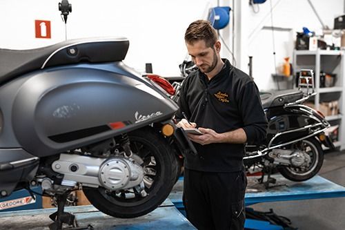 Een monteur inspecteert een grijze Vespa-scooter in een garage en maakt aantekeningen.