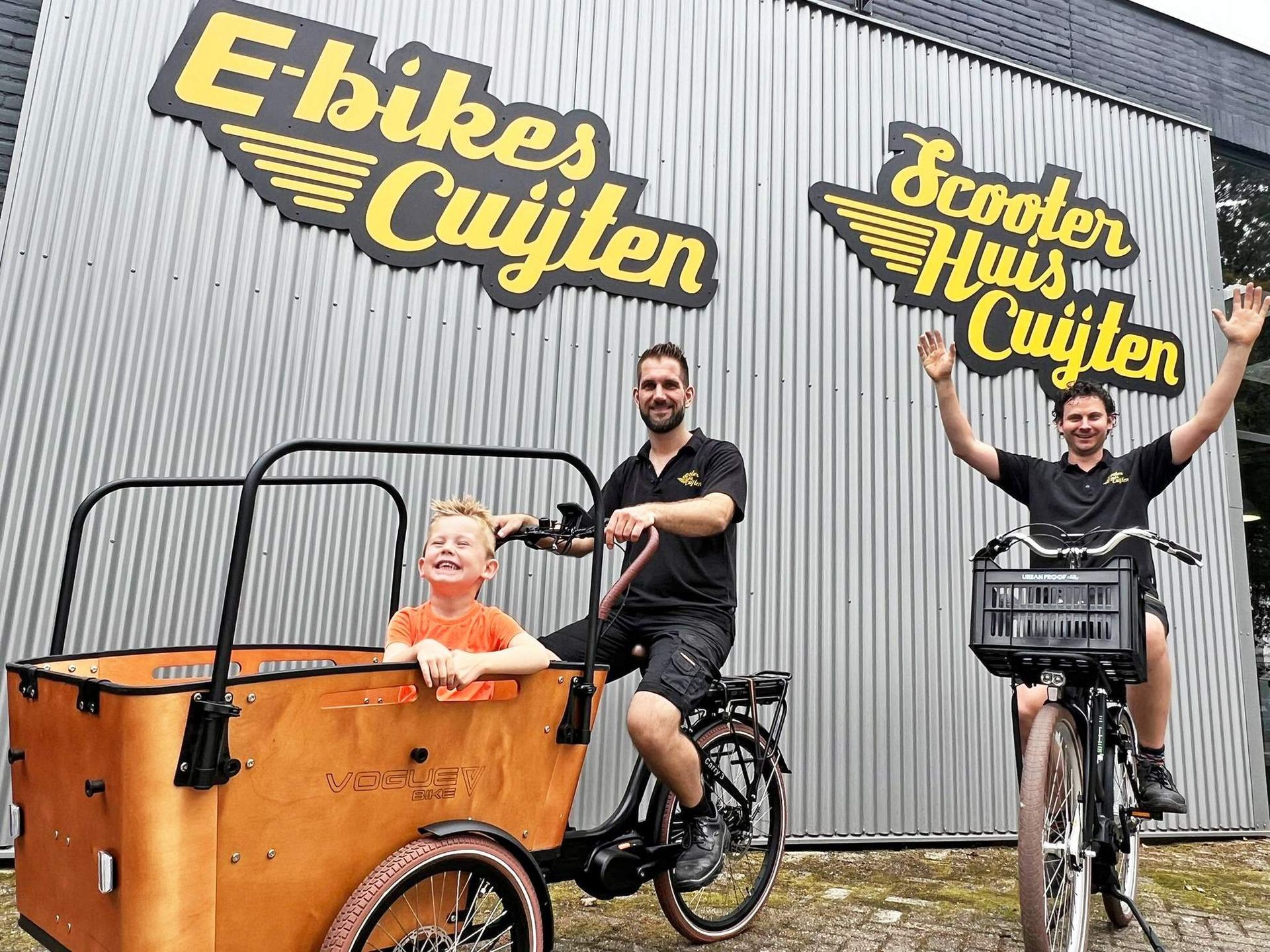 Mensen met e-bikes en een kind op een houten bakfiets poseren voor een winkel met reclameborden.
