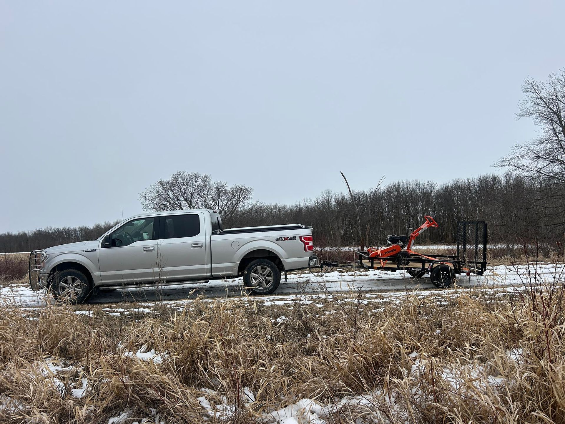 Silver pickup truck towing a red trailer through a snowy field under an overcast sky