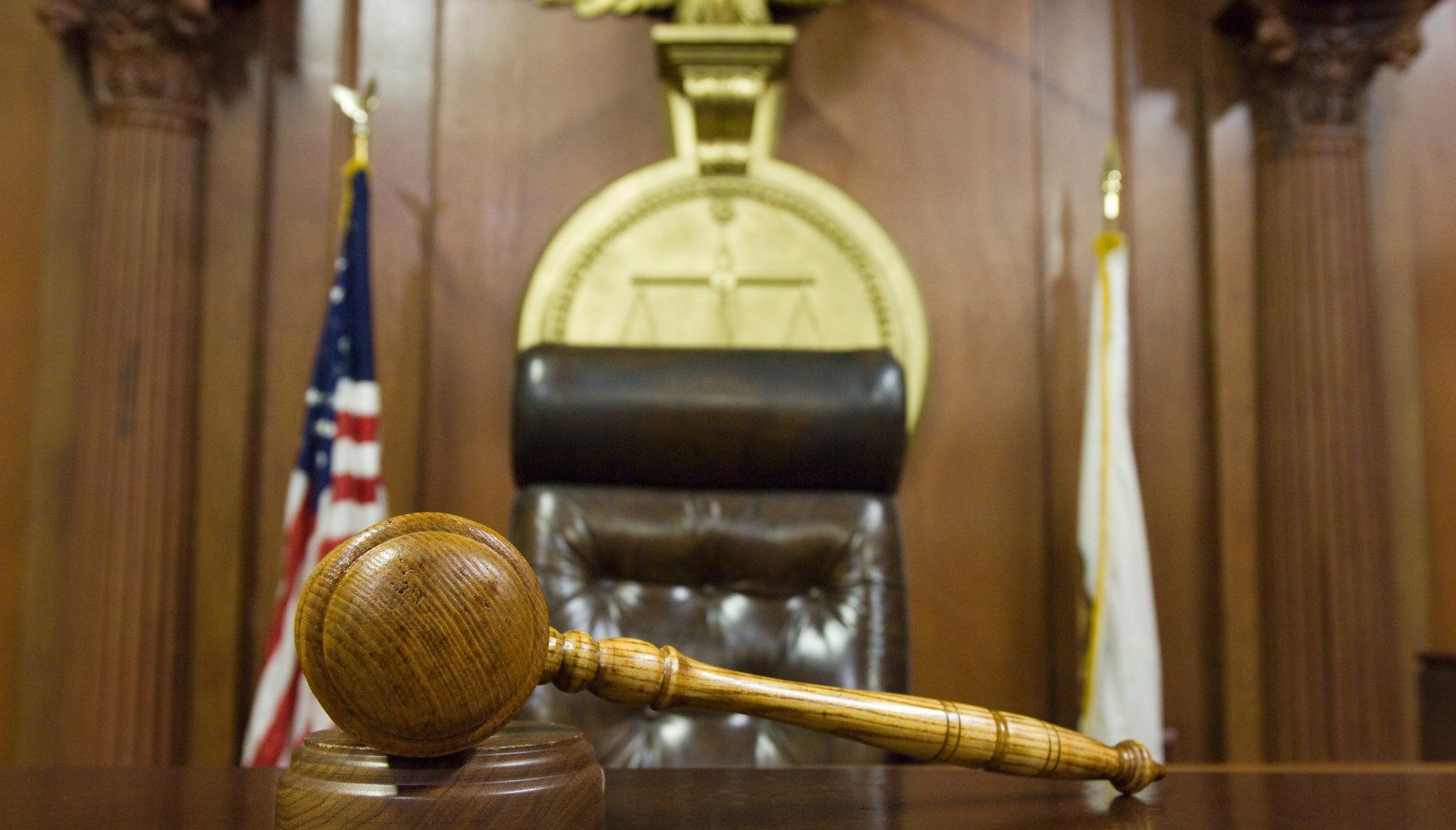 Gavel on a desk in a courtroom, with the judge's chair, scales, and flags in the background.