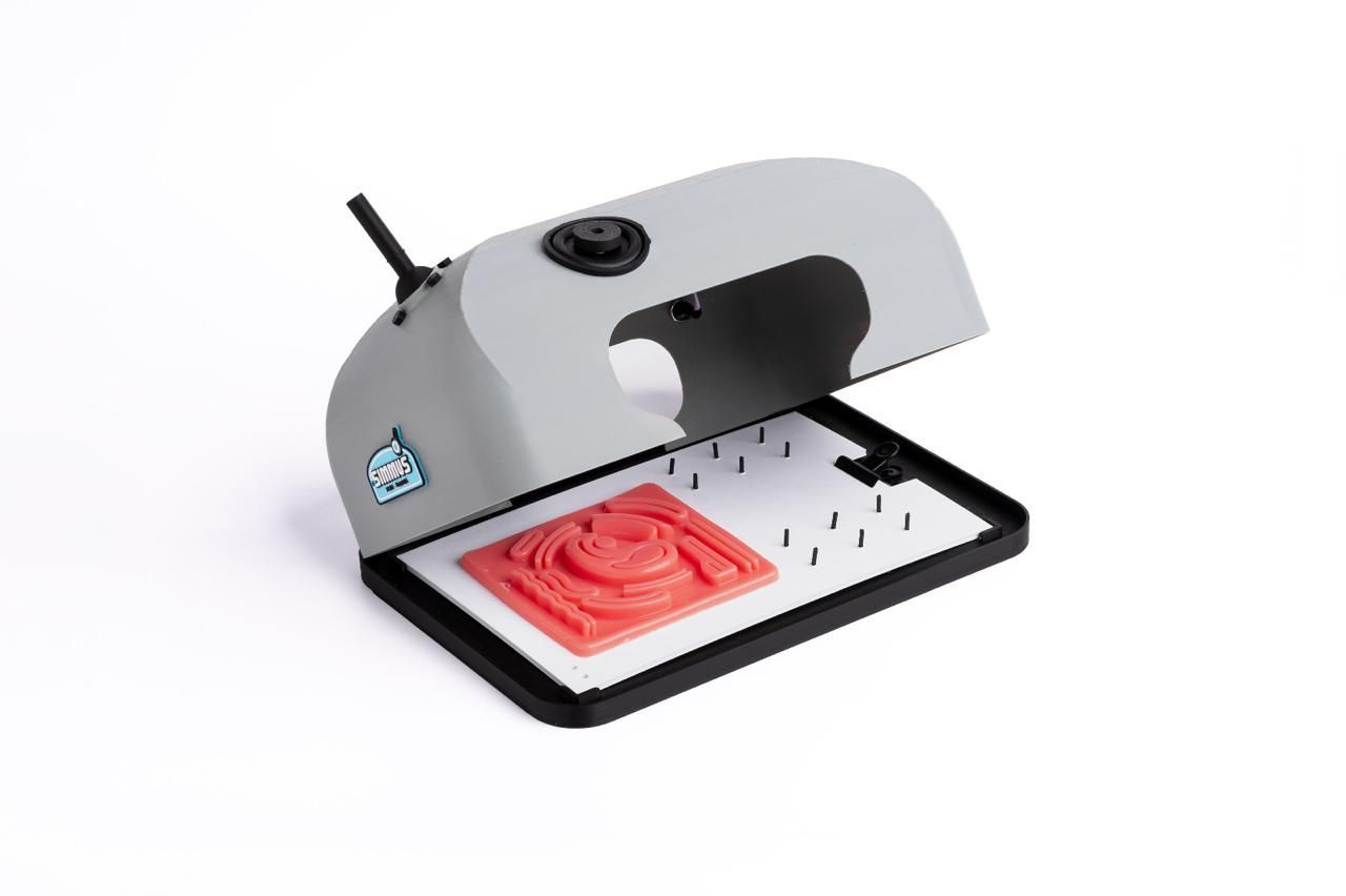 Unidad de luz ultravioleta gris abierta con un sello rojo y una bandeja blanca, probablemente para la fabricación de sellos.